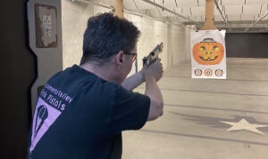 LGBTQ target shooter wearing a Pink Pistols t-shirt firing a handgun at a pumpkin target in an indoor shooting range