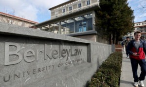 Photo a college student walking by a concrete sign with large silver lettering that reads, "Berkeley Law, University of California"