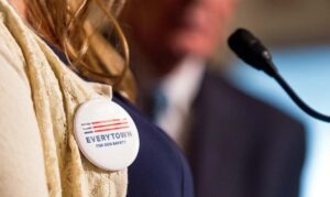 A close up photo of a large round pin reading "Everytown for gun safety" pinned to the white sweater of a woman with blond hair speaking into a small podium microphone. Article title: Everytown Comes Out From Behind the Curtain